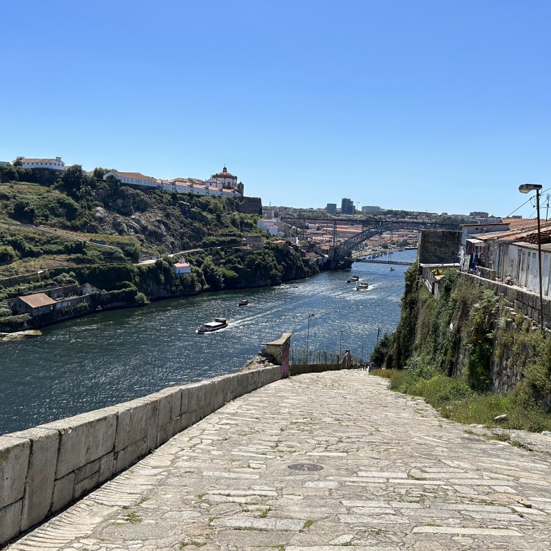 Riverside walkway with boats on water, sunny sky, and distant arch bridge.