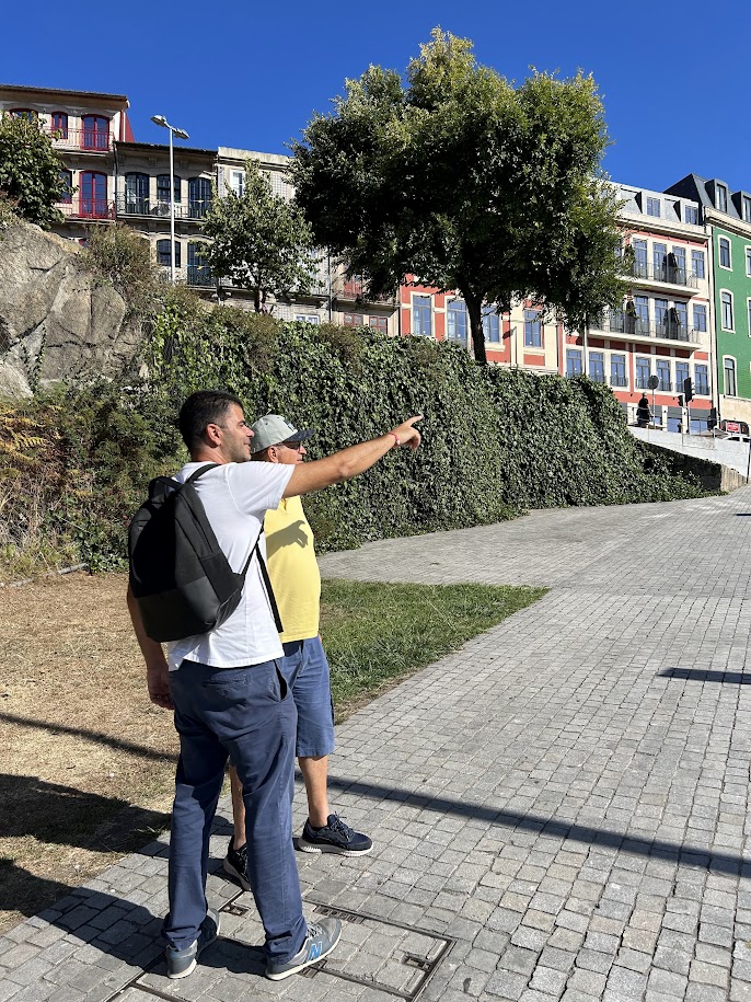 Two men on a street with one pointing, buildings and trees in the background.